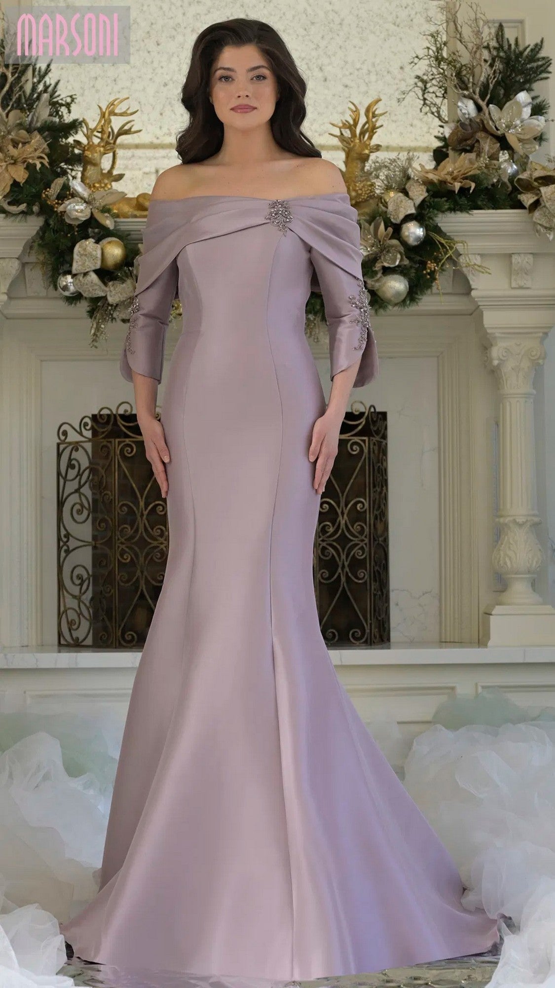 A woman stands indoors wearing the Marsoni MV1360 long formal dress in dusty rose, featuring a floor-length, off-the-shoulder design with a fitted bodice and brooch detail. Festive decorations are visible in the background.